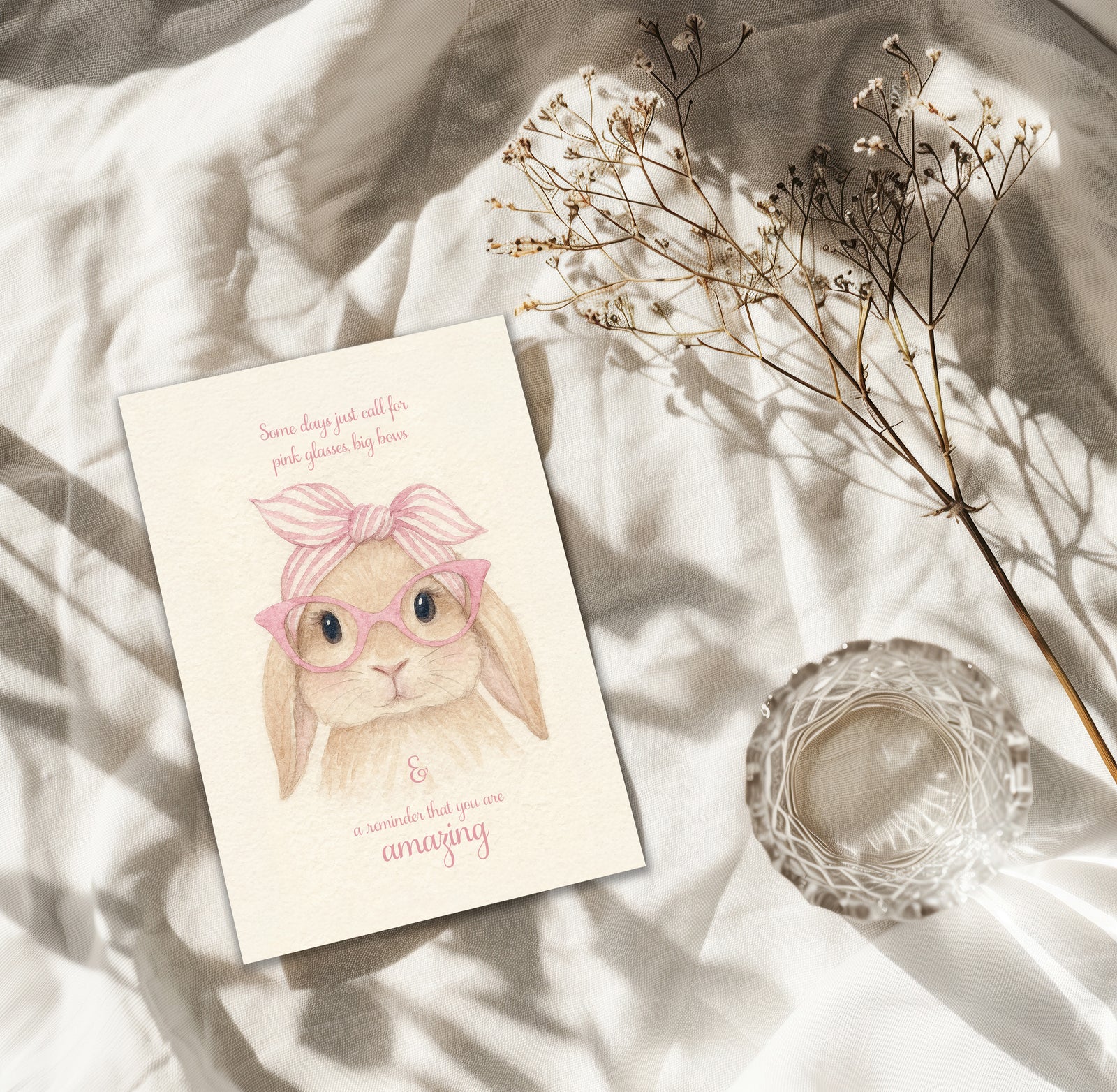 Card with a bunny illustration wearing glasses on a soft surface with dried flowers and a candle.